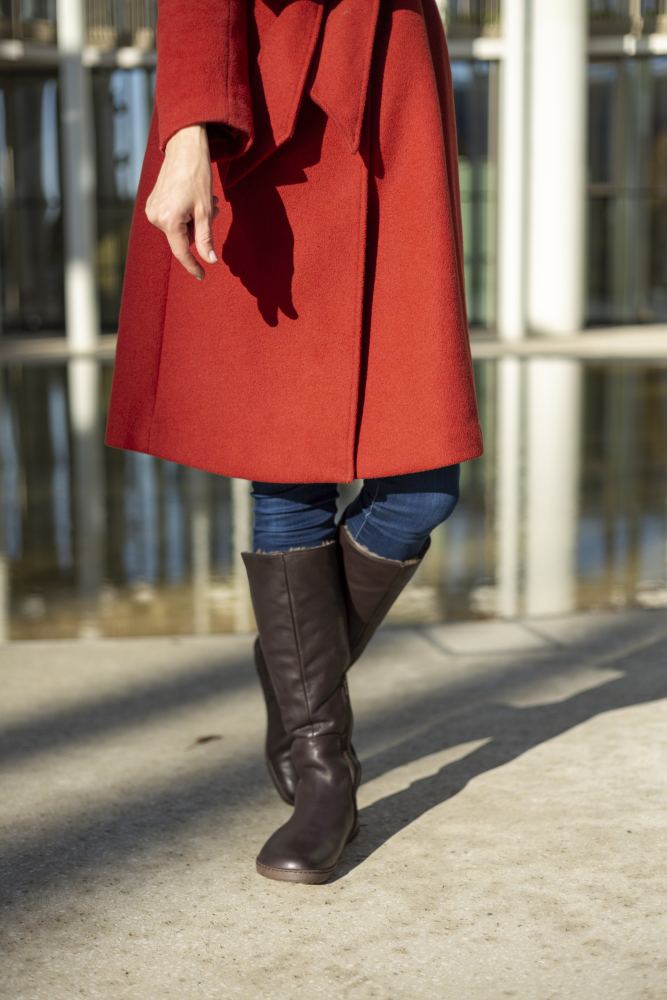 Eine Person in einem roten Mantel, Blue Jeans und ZAQQs kniehohen Stiefeln QATCH Winter Brown mit Lammfell-Futter spaziert auf einem sonnenbeschienenen Außenweg. Der Hintergrund besteht aus reflektierendem Glas mit vertikalen Linien.