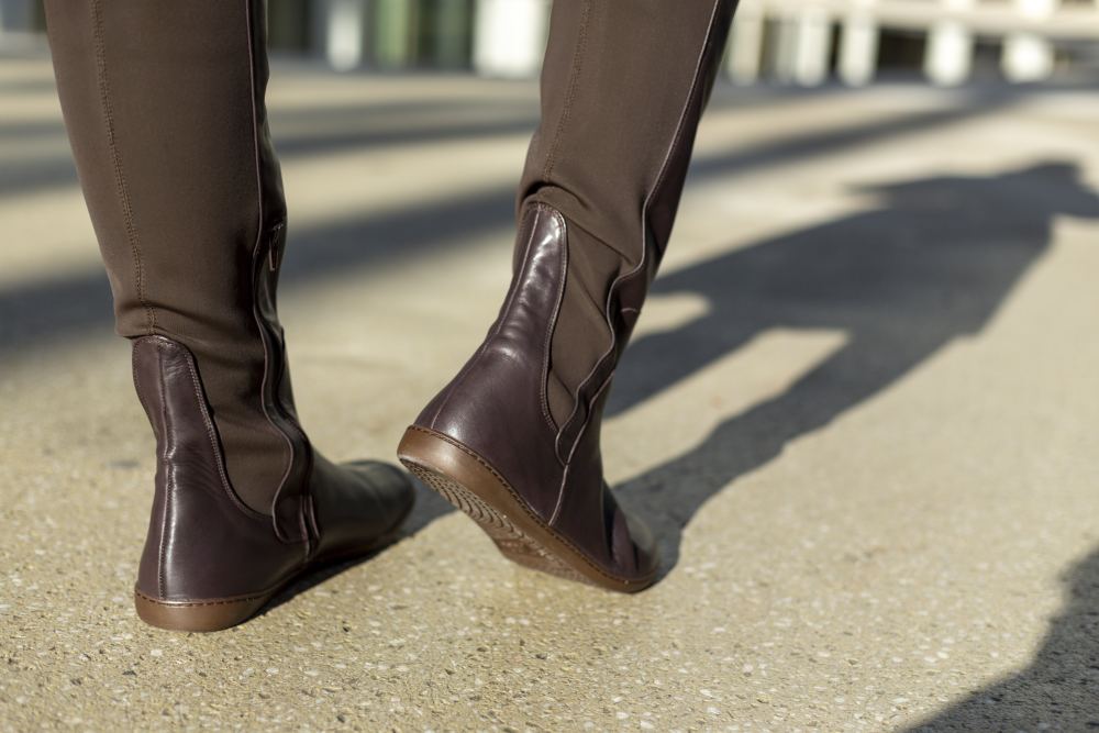 Eine Nahaufnahme von jemandem, der auf einem sonnenbeschienenen Bürgersteig spazieren geht und ZAQQs QATCH Nappa Brown-Stiefel und dunkle Hosen trägt. Die Betonung liegt auf dem Barfußschuh und den Schatten, die die Beine werfen.