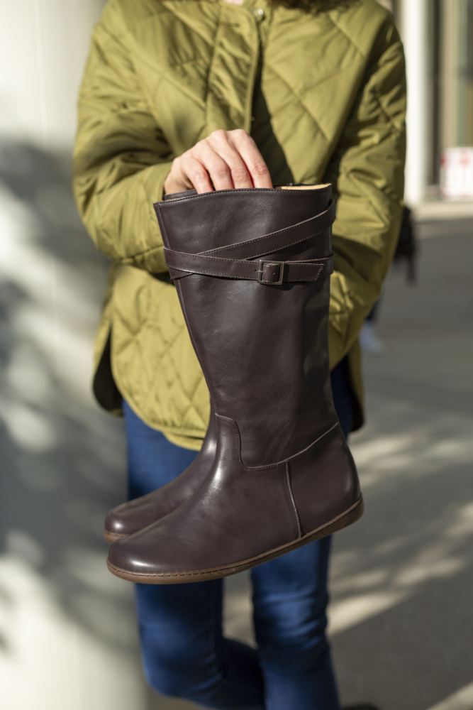 Eine Person in einer grünen Jacke hält ein Paar hohe, braune ATTRAQT Nappa Brown-Stiefel mit Riemen- und Schnallendesign. Diese aus luxuriösem Ziegenleder von ZAQQ gefertigten Stiefel werden in einer Outdoor-Umgebung elegant präsentiert.