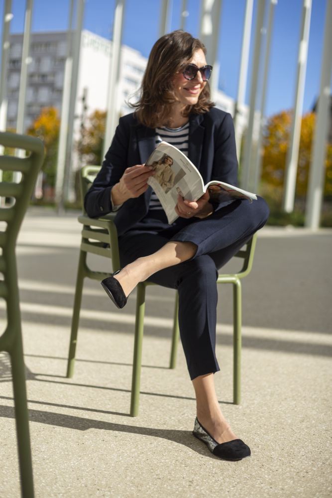 Eine Frau mit Sonnenbrille sitzt draußen auf einem grünen Stuhl und liest eine Zeitschrift. Sie trägt einen marineblauen Blazer und ein gestreiftes Hemd und kombiniert ihr Outfit mit schwarzen ZAQQ TWIST Derby-Schuhen. Sonnenlicht wirft Schatten auf den Boden und im Hintergrund sind Gebäude mit Herbstbäumen zu sehen.