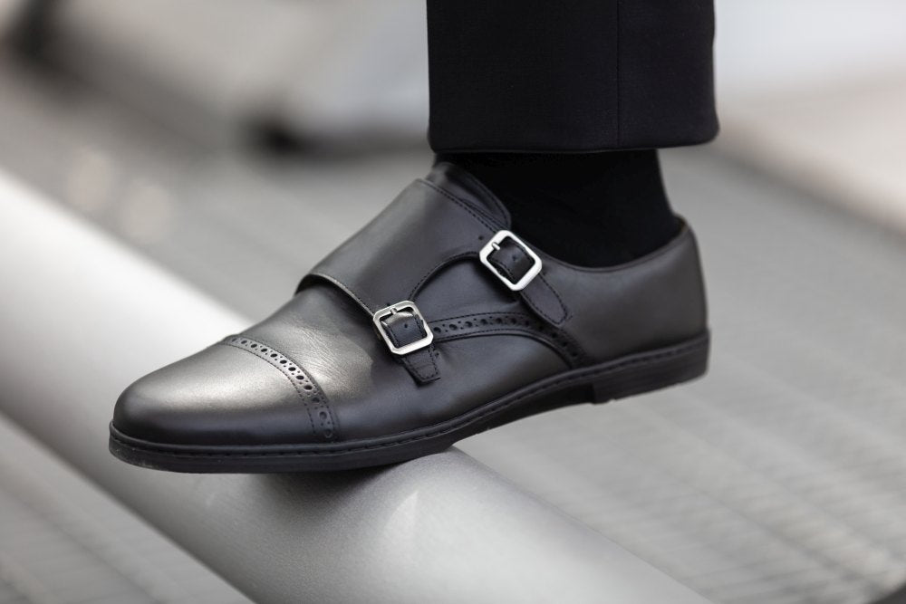 Eine Nahaufnahme des eleganten ZAQQ MONQ Captoe Black Schuhs, einem schwarzen Monkstrap-Businessschuh aus Leder mit zwei silbernen Schnallen, getragen von jemandem in stilvollen schwarzen Hosen. Der Schuh ruht selbstbewusst auf einer Metalloberfläche, seine Barfußsohle verleiht vor einem verschwommenen Hintergrund einen Hauch von Komfort.