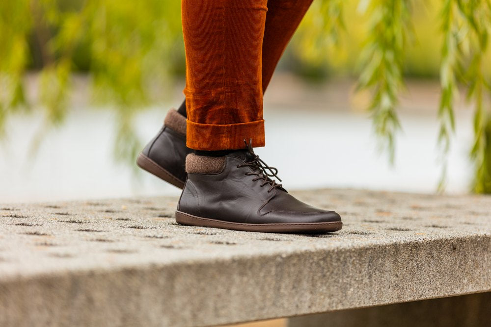 Eine Person in NEWQUAY Brown-Stiefeletten von ZAQQ und orangefarbenen Hosen steht auf einer Betonfläche. Der Hintergrund ist verschwommen, mit Andeutungen von Grün und Wasser.