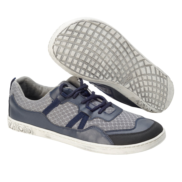 Ein Paar SQATE Grey-Sneaker von ZAQQ mit schicken grauen und blauen Mesh-Akzenten und weißen Sohlen. Ein Schuh ist seitlich positioniert, um das Gittermuster auf der Sohle zu zeigen, während der andere aufrecht steht, um die Schnürsenkel und das Obermaterialdesign hervorzuheben.