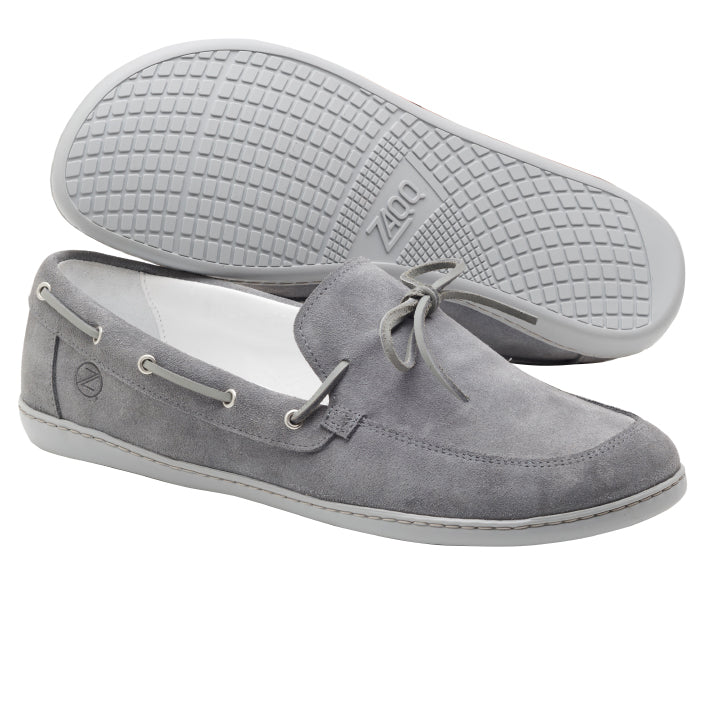 Ein Paar QEA Velours Grey Loafer im Mokassin-Stil von ZAQQ aus hochwertigem Veloursleder mit Gummisohle. Diese Barfußschuhe verfügen über eine dekorative Schnürung, ein dezentes Logo an der Seite und werden so präsentiert, dass ein Schuh aufrecht steht und der andere flach liegt, um die Sohle hervorzuheben.