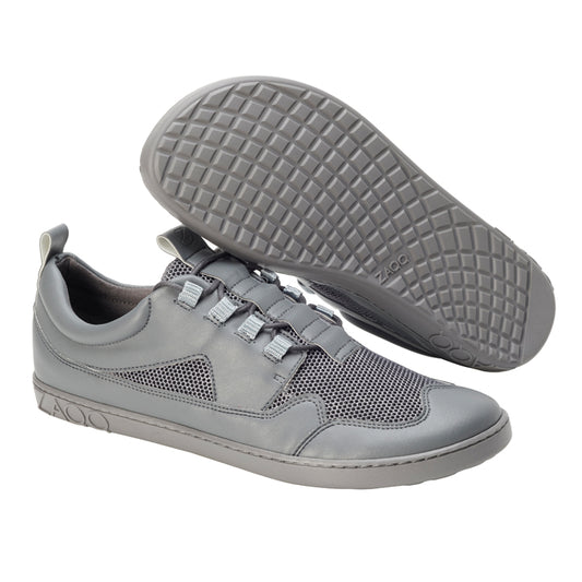 Ein Paar QARLEE Grey-Sneaker von ZAQQ, bei denen ein Schuh aufrecht steht und der andere auf der Seite liegt, hebt eine strukturierte Gummisohle hervor. Das Design umfasst einen Mesh-Einsatz und verstellbare Schnürsenkel und sorgt für ein modernes und sportliches Aussehen.