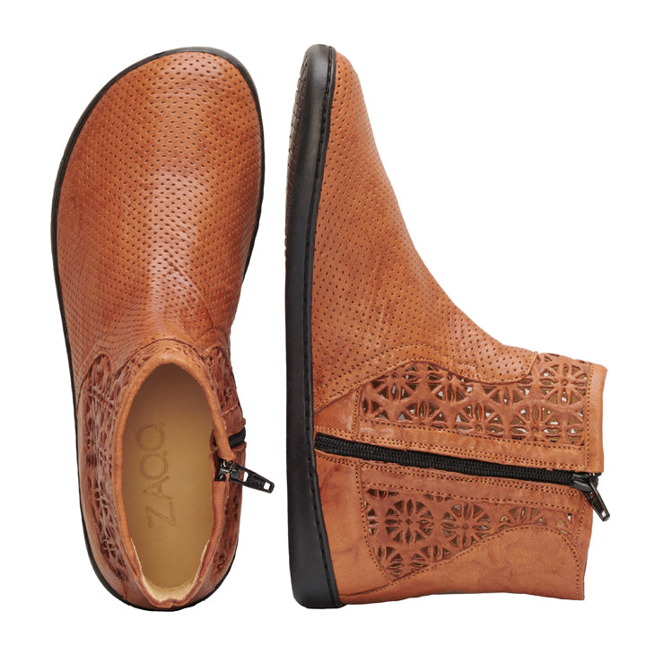 Ein Paar QYLIE Cognac-Lederstiefeletten von ZAQQ mit dekorativen Perforationen und seitlichen Reißverschlüssen. Ein Stiefel ist im Profil zu sehen, wodurch die aufwendigen Ausschnittdetails hervorgehoben werden, während der andere Stiefel von oben betrachtet wird und den Markennamen ZAQQ auf der Innensohle zeigt. Diese vielseitigen Barfußstiefel sind ideal für jeden Anlass.