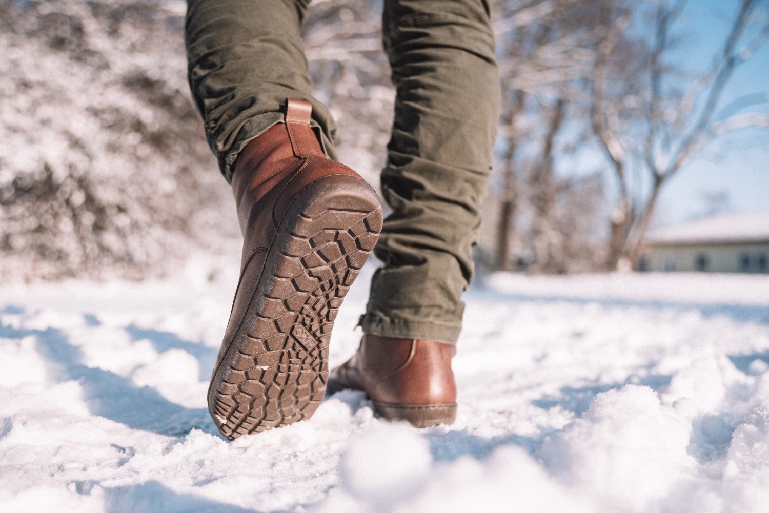 Eine Person in braunen Lederstiefeln und grünen Hosen läuft durch den Schnee. Die Sohlen der Stiefel haben ein strukturiertes Muster. Im Hintergrund sind kahle Winterbäume unter einem klaren blauen Himmel zu sehen, die eine ruhige Winterszene schaffen.