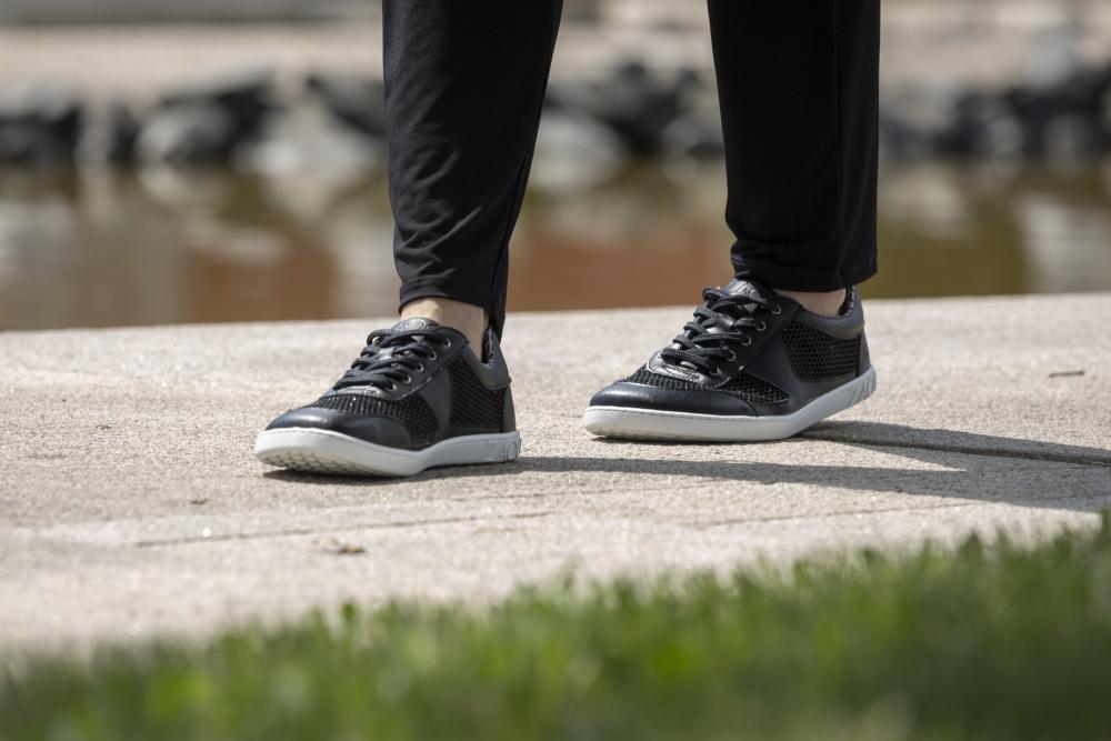 Eine Person schreitet einen Betonweg entlang und trägt schwarze ALIQANTE-Sneaker mit weißer Sohle von ZAQQ. Das verschwommene Grün und eine schimmernde Wasserfläche bilden den Hintergrund, während die schwarze Hose den Look vervollständigt.