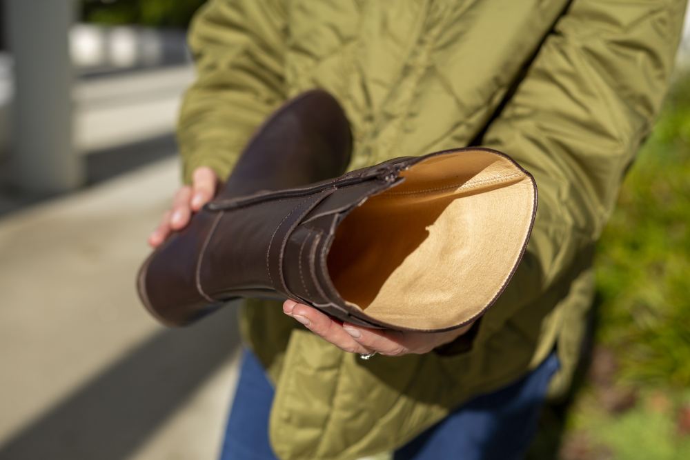 Jemand präsentiert stolz einen veganen braunen ATTRAQT-Stiefel von ZAQQ und zeigt sein Innenfutter. Die Person trägt eine grüne Steppjacke und blaue Jeans vor einem verschwommenen Outdoor-Hintergrund, der die Aufmerksamkeit auf die flexible Sohle und die Barfußschuh-Eigenschaften des Stiefels lenkt – ideal für Naturliebhaber, die sowohl Stil als auch Komfort schätzen.