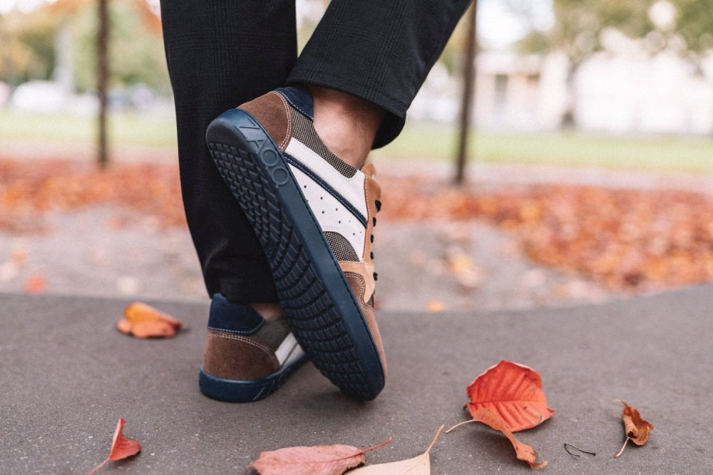 Nahaufnahme einer Person, die schicke braun-blaue BREAQ Sierra-Sneaker von ZAQQ mit weißen Akzenten trägt. Die Person steht auf einem mit Herbstblättern bedeckten Weg und trägt dunkle Hosen. Dahinter verstärken verschwommenes Grün und Bäume den Scheinwerfer auf die flexible Sohle des Sneakers.