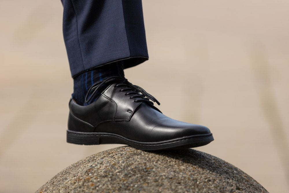 Eine Person in marineblauen Hosen und gestreiften Socken tritt in polierten BRIQ Black-Schuhen von ZAQQ auf einen runden Stein und sorgt so für einen stilvollen und modernen Auftritt. Das zeitlose Design dieser eleganten Barfußschuhe verleiht dem weichen, verschwommenen beigen Hintergrund Raffinesse.