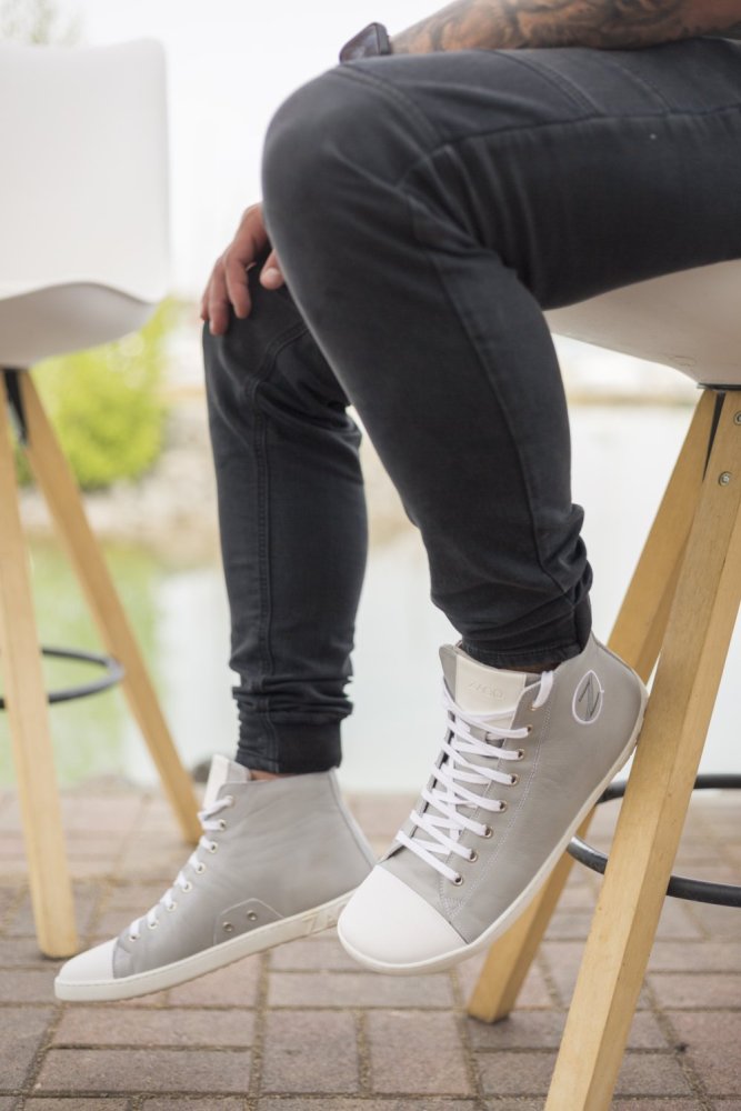 Eine Person sitzt auf einem Holzhocker und trägt ZAQQs CHUQQS Grey High-Top-Sneaker aus Nappaleder, die ihre schwarzen Jeans perfekt ergänzen. Der Hintergrund zeigt eine verschwommene Außenszene mit Grün und Wasser, die die ruhige Atmosphäre verstärkt.