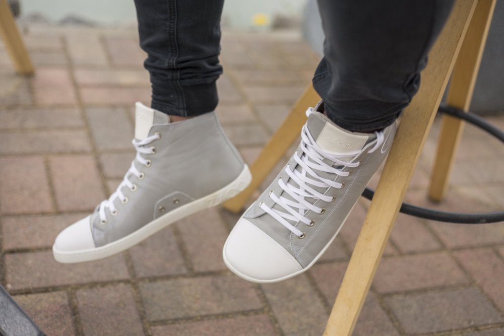 Nahaufnahme einer Person, die ZAQQs graue High-Top-Sneakers CHUQQS aus Nappaleder mit weißen Schnürsenkeln trägt und auf einem Holzhocker sitzt. Die Person trägt dunkle Hosen vor einem gepflasterten Hintergrund.