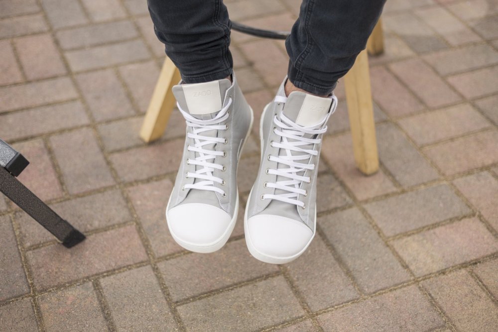 Eine Person trägt die grauen High-Top-Sneakers „CHUQQS“ von ZAQQ mit weißen Schnürsenkeln aus Nappaleder und sitzt auf einem Holzstuhl auf einem gepflasterten Platz.