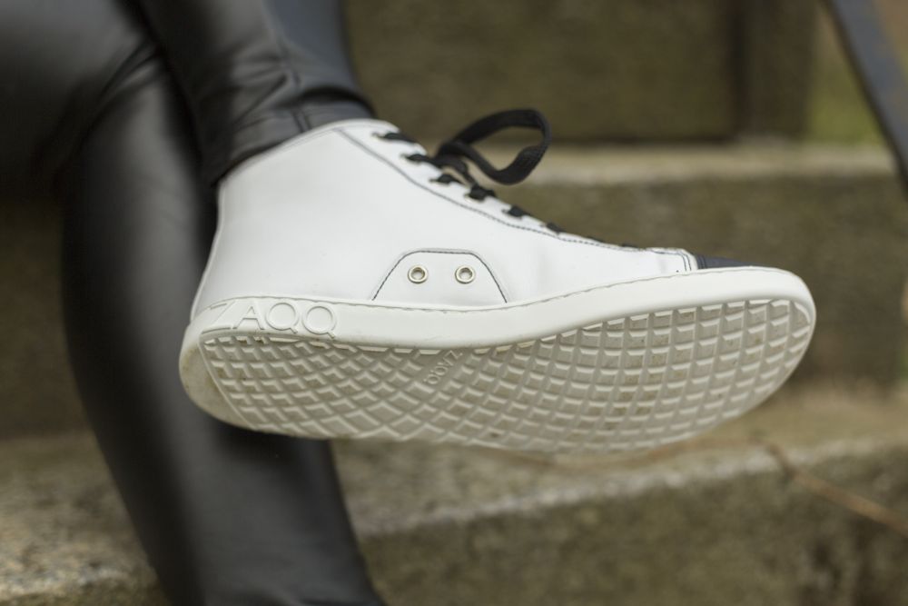 Eine Nahaufnahme des CHUQQS White-Sneakers von ZAQQ mit schwarzen Schnürsenkeln, markant strukturierten Sohlen und einem sichtbaren AQO-Logo. Die Person, die ihn trägt, sitzt auf Betonstufen und trägt schwarze Nappalederhosen.