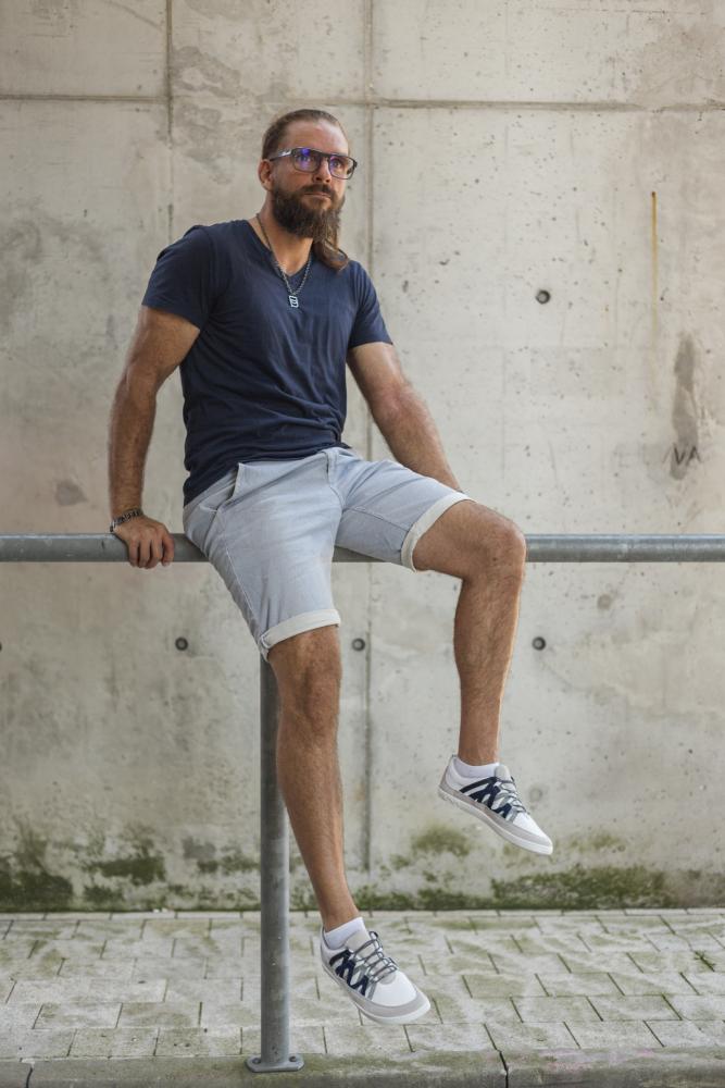 Ein Mann mit Bart und Brille sitzt lässig auf einem Geländer an einer Betonwand. Er trägt ein marineblaues T-Shirt, hellgraue Shorts und auffällige ZAQQ DRAQE White Grey Sneakers mit Akzenten aus hochwertigem Nappaleder.