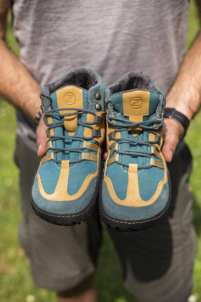 Eine Person hält draußen ein Paar blau-orangefarbene Wanderstiefel, im Hintergrund ist Gras zu sehen. Die wasserdichten Stiefel ZAQQ ESQAPE in Blau und Orange haben Schnürsenkel und dicke Sohlen, die für Komfort auf jedem Gelände sorgen.