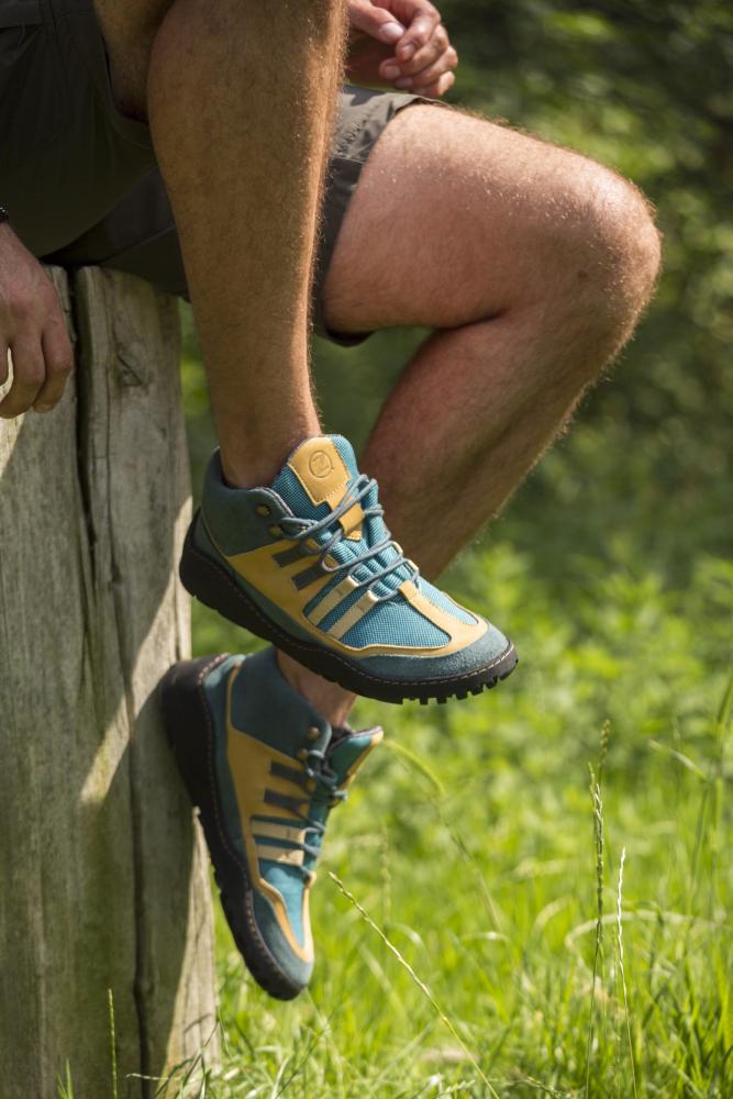 Eine Person in blauen und orangefarbenen wasserdichten Wanderschuhen von ZAQQ sitzt in Shorts auf einem Holzstumpf in einer sonnenbeschienenen Grasfläche.