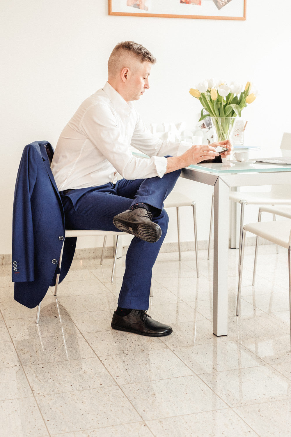 Ein Mann in weißem Hemd und blauer Hose sitzt mit übereinandergeschlagenen Beinen an einem Glastisch und konzentriert sich auf sein Telefon. Sein PEAQ Black von ZAQQ verleiht ihm einen Hauch von Eleganz, während eine blaue Jacke über dem Stuhl hängt. Eine Vase mit weißen und gelben Tulpen schmückt den Tisch in diesem hellen Raum.