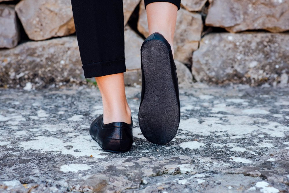 Eine Person in schwarzer Hose und einem Paar ZAQQ IQON Black Schuhen läuft auf einem Steinweg. Der Fokus liegt auf den Sohlen der Schuhe, die im klassischen Derby-Stil gefertigt sind, während sie vorwärts geht, mit einer Steinmauer im Hintergrund.