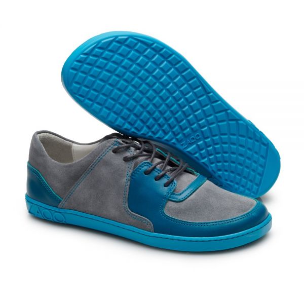 Ein Paar IVIQ Turquoise-Sneaker von ZAQQ mit einer blauen Gummisohle und einem Obermaterial aus grauem Wildleder und blauem Leder. Ein Schuh steht aufrecht, während der andere darüber zu sehen ist, wodurch die strukturierte Sohle hervorgehoben wird. Das Design umfasst Schnürsenkel und Nähte, die für hohen Komfort sorgen.