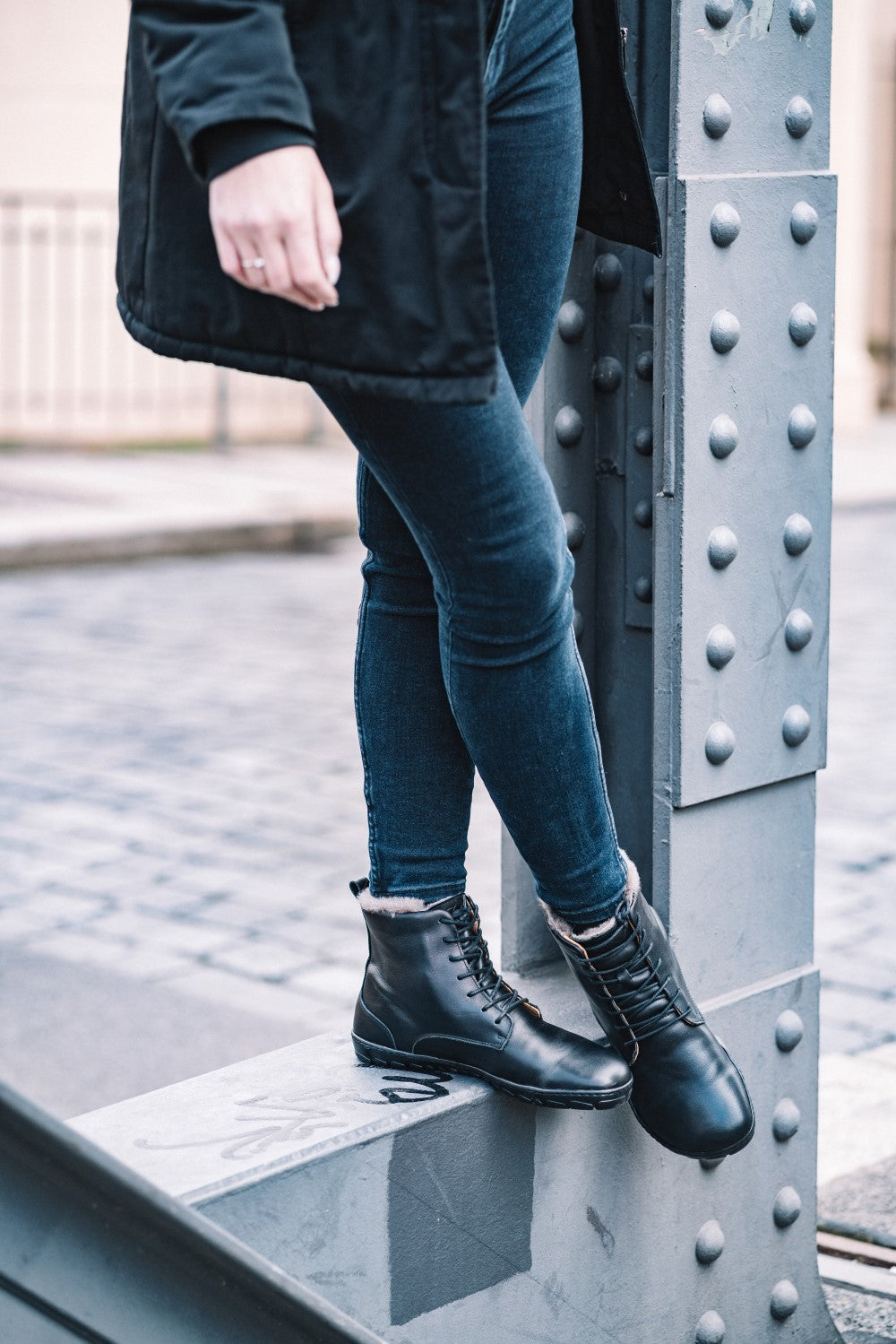 Eine Person in schwarzem Mantel und Röhrenjeans lehnt an einem genieteten Metallträger. Sie trägt die QUINTIC Winter Black Stiefel von ZAQQ, die perfekt für den kühlen Tag geeignet sind, und steht auf einer gepflasterten Stadtstraße.