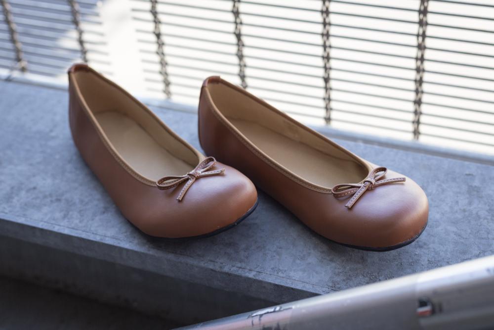 Ein Paar ZAQQ LOOQ Vegan Cognac Ballerinas mit kleinen Schleifen ruht auf einem Betonsims. Diese Ballerinas aus atmungsaktivem Material verfügen über eine Barfußsohle für ultimativen Komfort. Als Hintergrund dient ein Metallgeländer mit vertikalen Stäben.