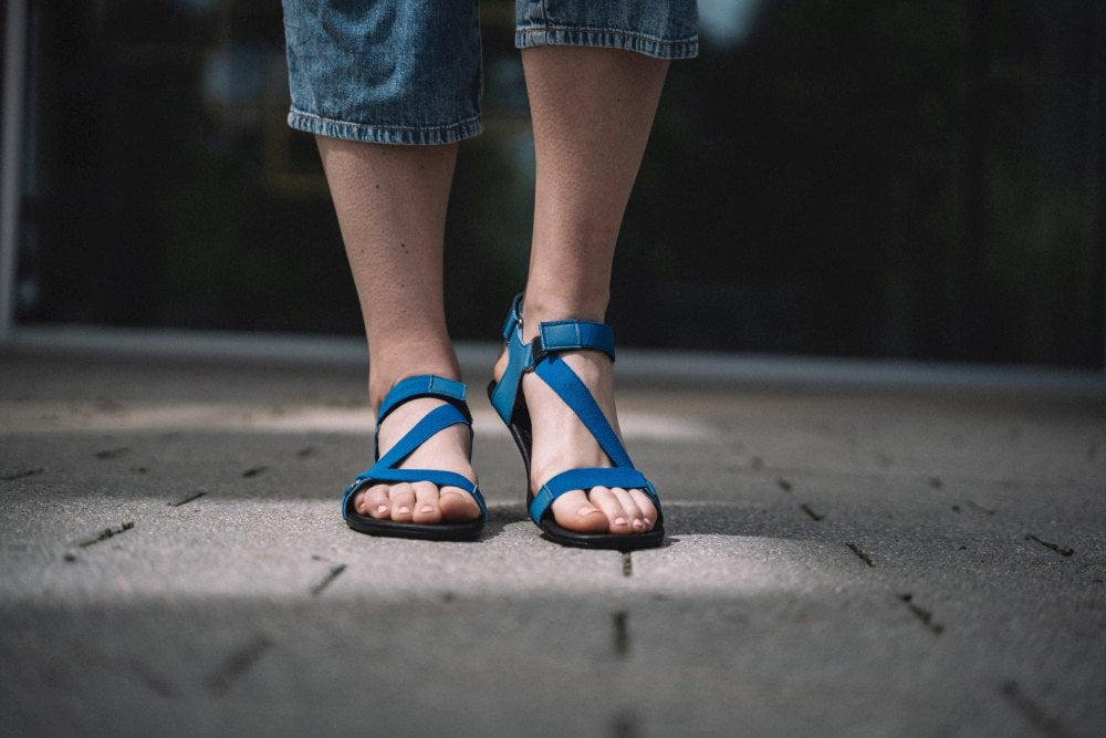 Nahaufnahme einer Person, die blaue MIQA Blue-Sandalen von ZAQQ mit Gummisohlen trägt und auf einem strukturierten Bürgersteig steht. Die verkürzten Jeans heben das Schuhwerk hervor und der Fokus liegt auf den Füßen. Der unscharfe Hintergrund verleiht der Szene Tiefe.