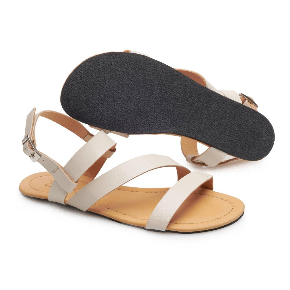 Wir präsentieren die NIQ Beige Sandalen von ZAQQ, ein Paar flache Damensandalen aus hochwertigem Nappaleder. Sie zeichnen sich durch kontrastierende hellbraune Sohlen und elegante weiße Riemen aus. Eine Sandale zeigt die schwarze strukturierte Unterseite, während die andere eine Ansicht von oben bietet. Der Knöchelriemen ist mit einer Metallschnalle akzentuiert, wodurch diese Sandalen ideal zu einem luftigen Sommerkleid passen.