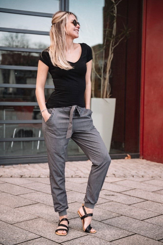 Eine Frau mit NIQ Black-Sonnenbrille und schwarzem T-Shirt, dazu graue Hosen und moderne ZAQQ-Sandalen, steht lächelnd vor einem Gebäude, die Hände in den Taschen. Ein großes Fenster und eine rote Wand bilden den Hintergrund und strahlen eine Aura von maximalem Komfort aus.