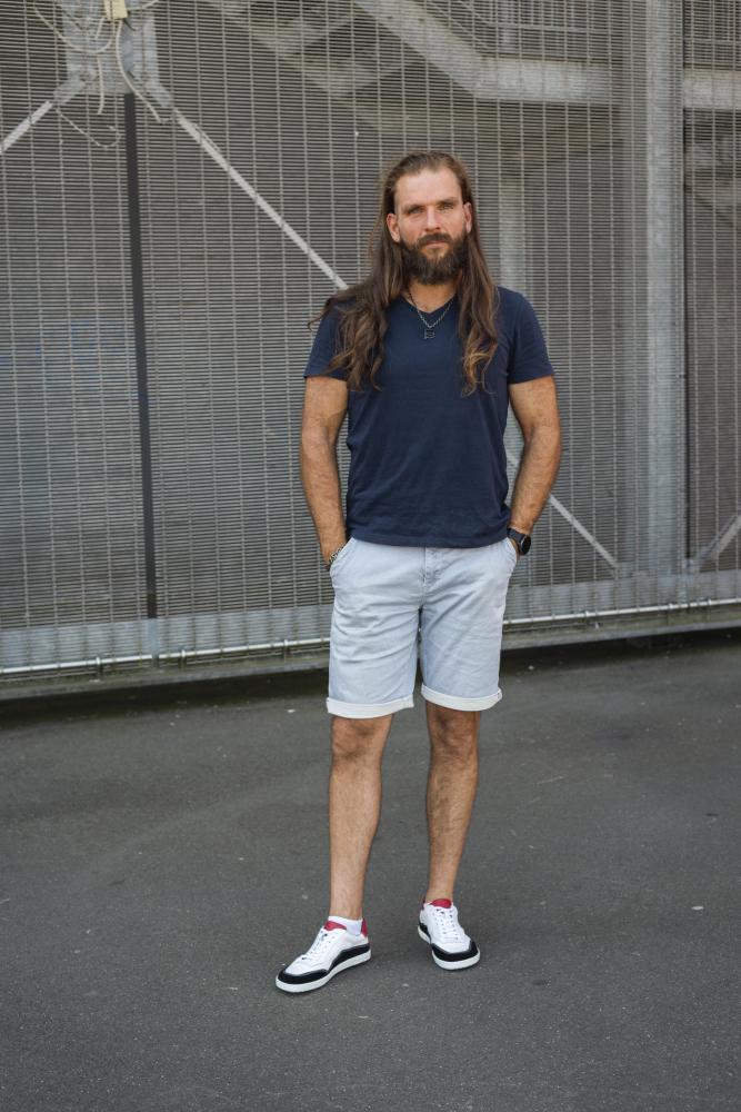 Ein Mann mit langen Haaren und Bart steht mit neutralem Gesichtsausdruck im Freien. Er trägt ein dunkelblaues T-Shirt, hellgraue Shorts und weiße QAMPION Black White-Sneaker von ZAQQ mit roten Details und flexibler Sohle. Im Hintergrund sind ein Metallgitterzaun und eine dunkle Gehwegoberfläche zu sehen.