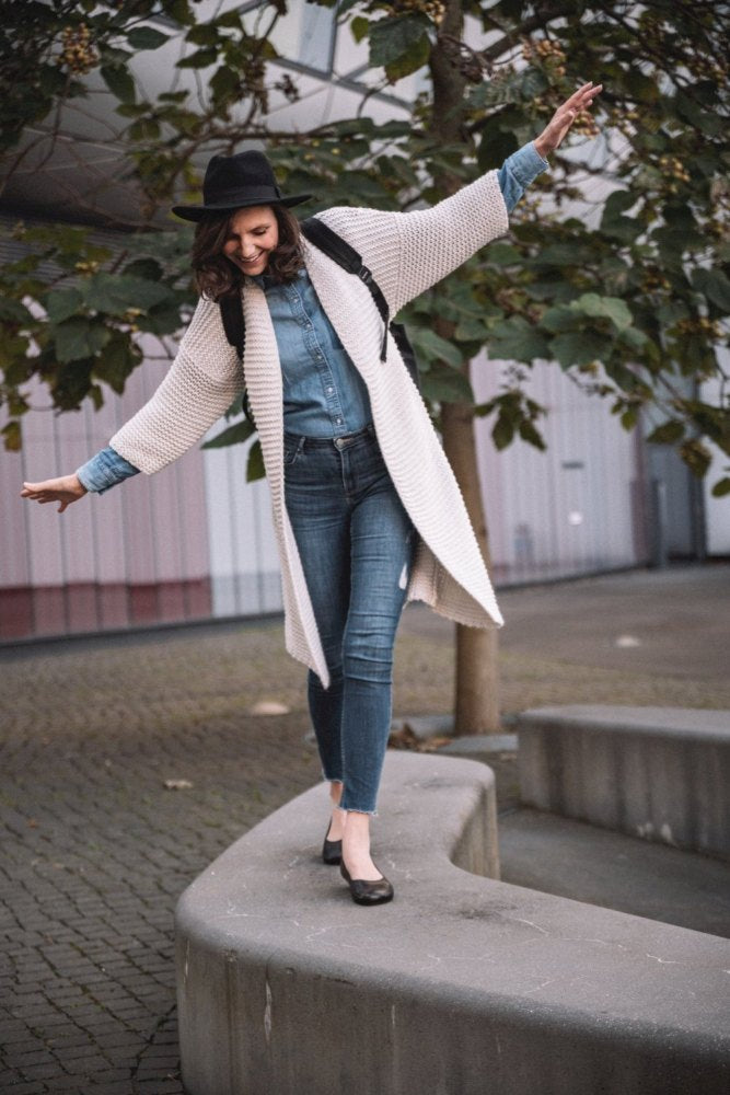 In einer stylischen Strickjacke aus Nappaleder, Jeans und einem Hut balanciert eine Person spielerisch auf einem geschwungenen Betonvorsprung. Sie trägt einen dunkelbraunen ZAQQ QARMA-Rucksack und genießt die Natur in der Nähe einiger Bäume und eines modernen Gebäudes. Sie fängt den Moment mit der Anmut einer Barfuß-Ballerina ein.