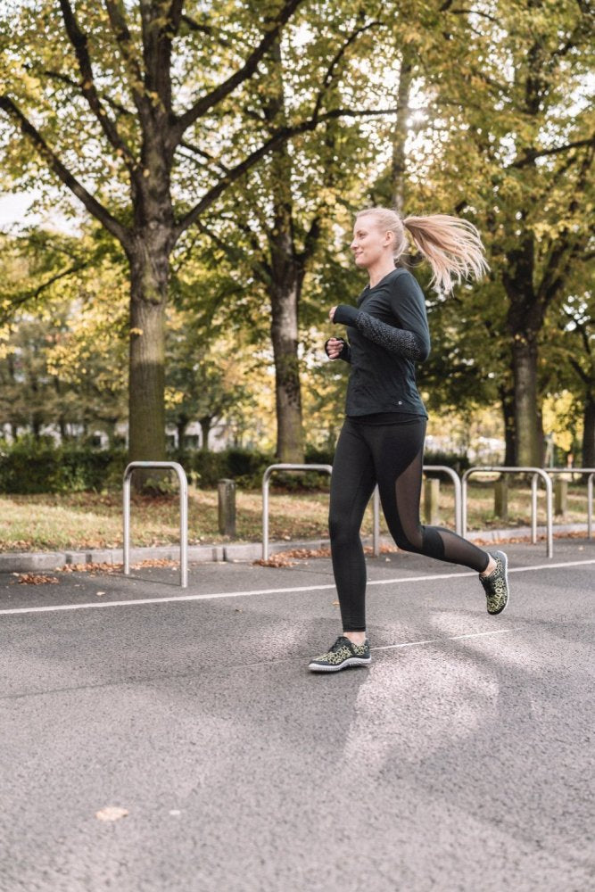 In Sportbekleidung und dem QASEY Green von ZAQQ läuft eine Person auf einem gepflasterten Weg im Park entlang, umgeben von hohen Bäumen mit üppigen grünen Blättern. Die helle, heitere Atmosphäre lässt darauf schließen, dass es ein sonniger Tag ist. Mit ihren zurückgebundenen langen Haaren wirkt sie auf ihren Lauf konzentriert und genießt den Komfort des atmungsaktiven Mesh-Materials.