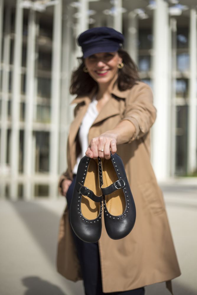 Eine Frau in einem beigen Trenchcoat und einer marineblauen Mütze hält ein Paar DIEQO Nappa Black-Schuhe von ZAQQ in die Kamera. Im Hintergrund ist ein modernes Gebäude mit vertikalen Säulen zu sehen.