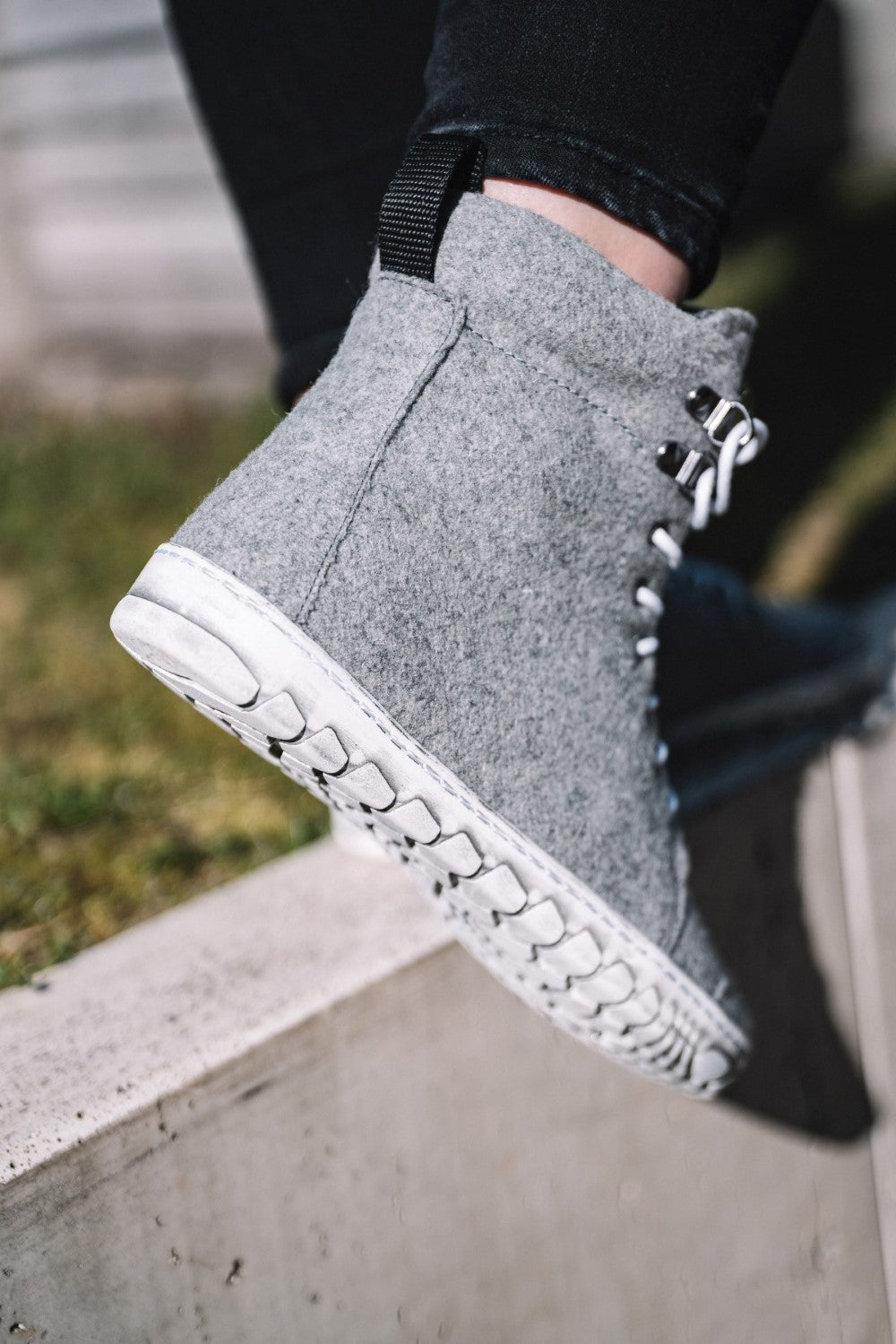 Nahaufnahme einer Person, die QICE Grey High-Top-Sneakers von ZAQQ aus Merinowolle mit weißen Sohlen und Schnürsenkeln trägt und auf einem Betonvorsprung steht. Die Schuhe haben eine flauschige Textur und die Person trägt eine schwarze Hose. Im unscharfen Hintergrund ist Gras zu sehen.