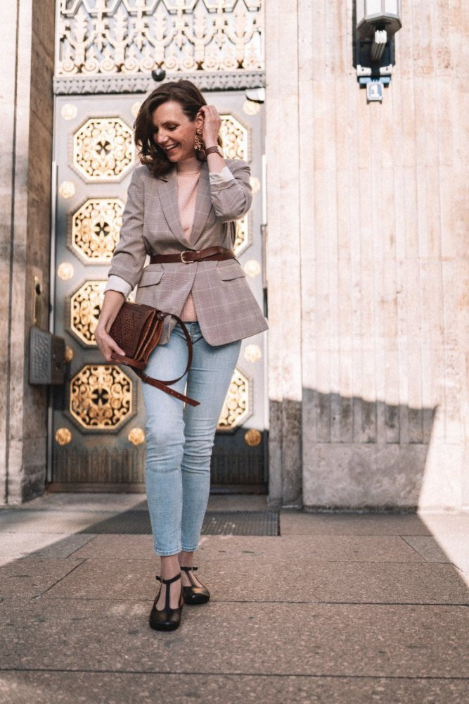 Eine Frau in kariertem Blazer und Jeans steht vor einem Gebäude mit verzierten goldenen Türen. Sie trägt eine dunkelbraune ZAQQ QITE-Handtasche, schwarze Ballerinas mit Barfußsohle und lächelt im Sonnenlicht.