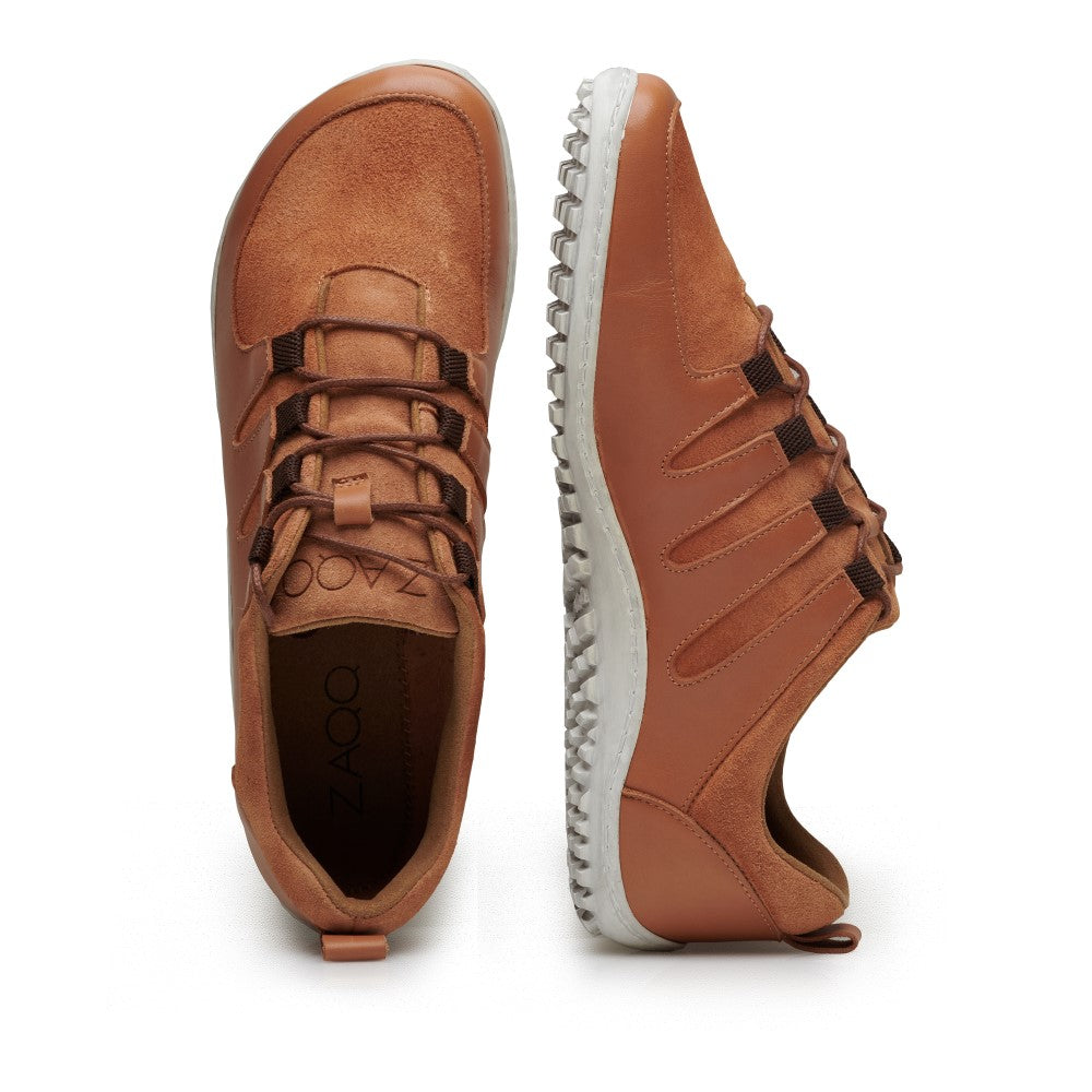 Zwei SHOQQ-Cognac-Sneaker von ZAQQ werden vorgestellt. Einer ist aufrecht positioniert, um die Draufsicht zu zeigen, während der andere auf der Seite liegt und das Design der strukturierten weißen Sohle zeigt. Diese Barfußschuhe haben Schnürfronten und ein sichtbares Branding im Inneren eines Schuhs, was ihren außergewöhnlichen Tragekomfort unterstreicht.