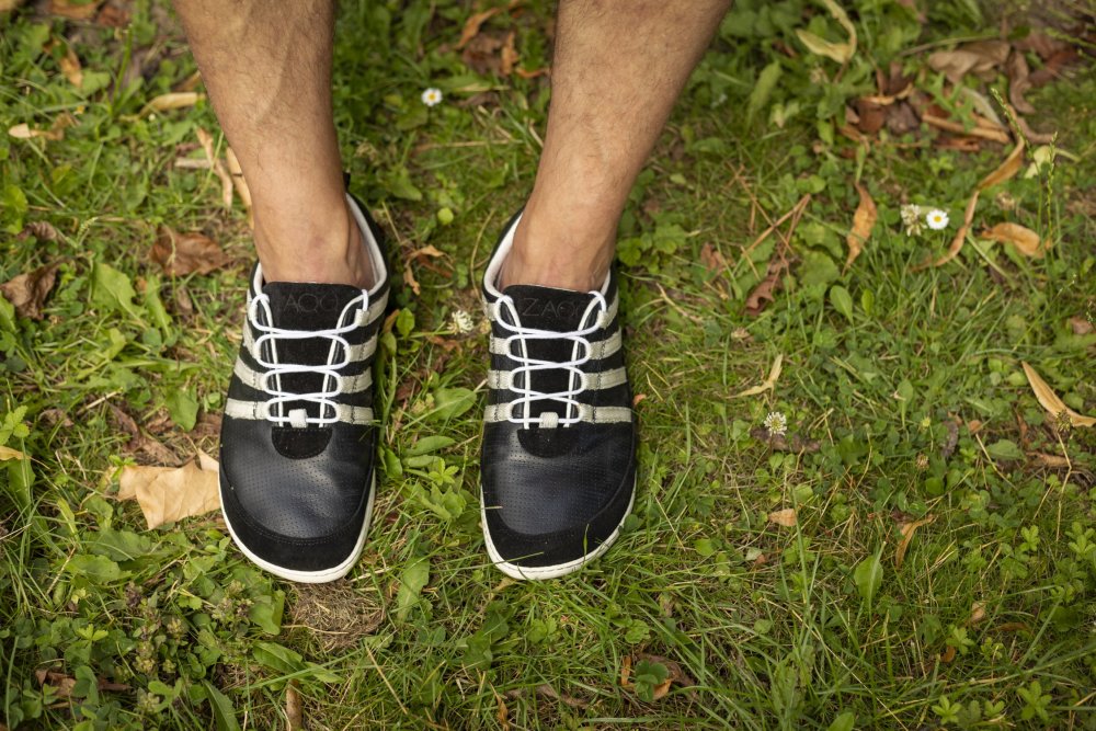 Nahaufnahme einer Person, die die SPARQ Low Black White-Sneakers von ZAQQ mit gestreiften Schnürsenkeln aus geschmeidigem Nappaleder trägt und auf Gras steht, umgeben von verstreuten Blättern und kleinen Blumen.