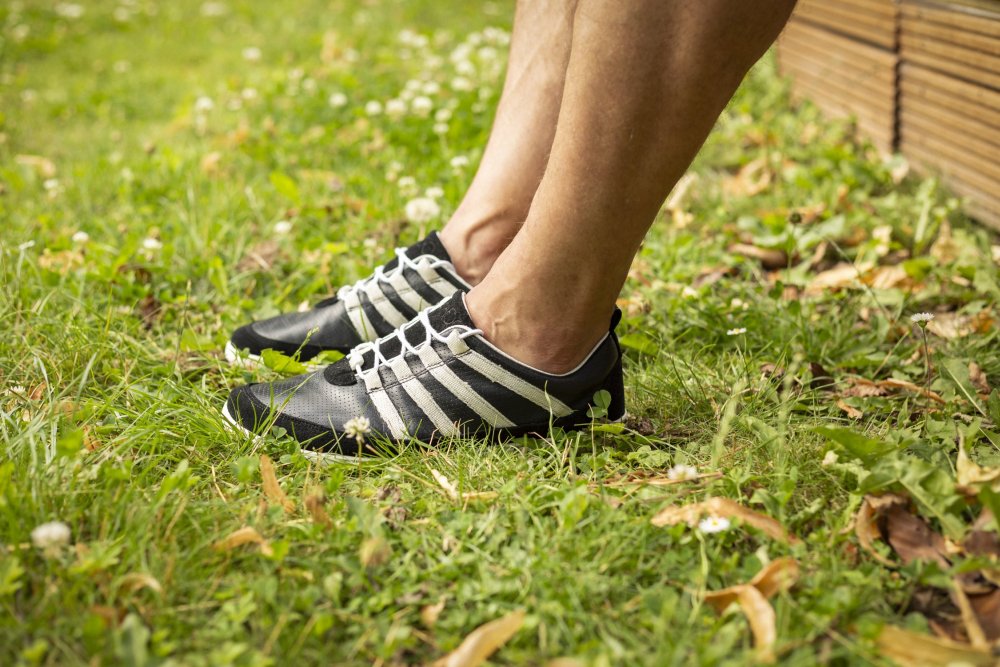 Eine Person steht auf dem Rasen und präsentiert die SPARQ Low Black White-Sneaker von ZAQQ. Das saftig grüne Gras ist mit kleinen weißen Blumen und abgefallenen Blättern übersät, im Hintergrund ist ein Holzzaun zu sehen.