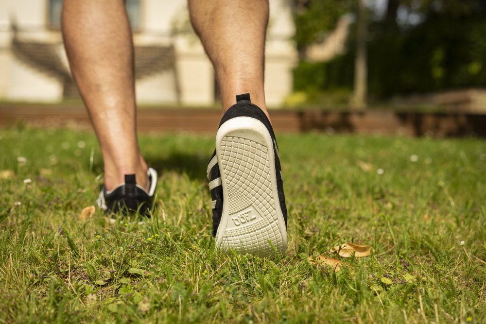 Eine Person läuft auf Gras und trägt die schicken ZAQQ SPARQ Low Black White Sneaker mit sichtbarer Sohle. Der Hintergrund ist verschwommen und zeigt ein Gebäude und Grünflächen unter einem sonnigen Himmel.