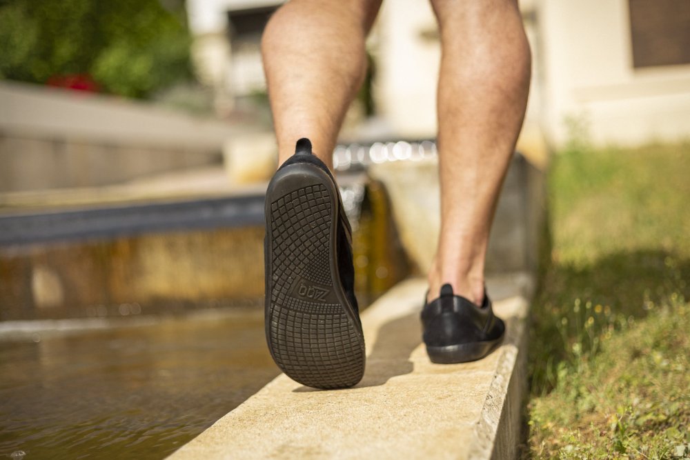 Nahaufnahme der Beine einer Person, die die eleganten SPARQ Low Black-Sneaker von ZAQQ mit schwarzer Sohle trägt, während sie am Rand eines seichten Wassers in einer Gartenlandschaft entlangläuft. Sonnenlicht wirft Schatten und hebt die strukturierte Sohle der Schuhe hervor.