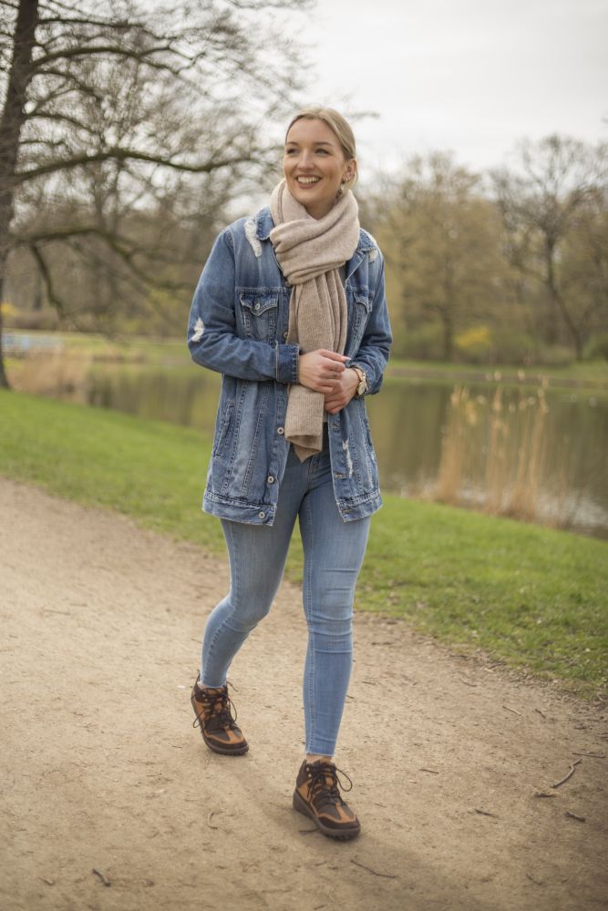 Eine Frau schlendert einen Kiesweg durch einen Park. Sie trägt eine Jeansjacke, Jeans und die ZAQQ SQOUT Brown Waterproof Wanderschuhe mit der strapazierfähigen TERRA-Sohle. Ihr beigefarbener Schal flattert, während sie lächelt. Ein Teich und Bäume bieten die perfekte Kulisse.