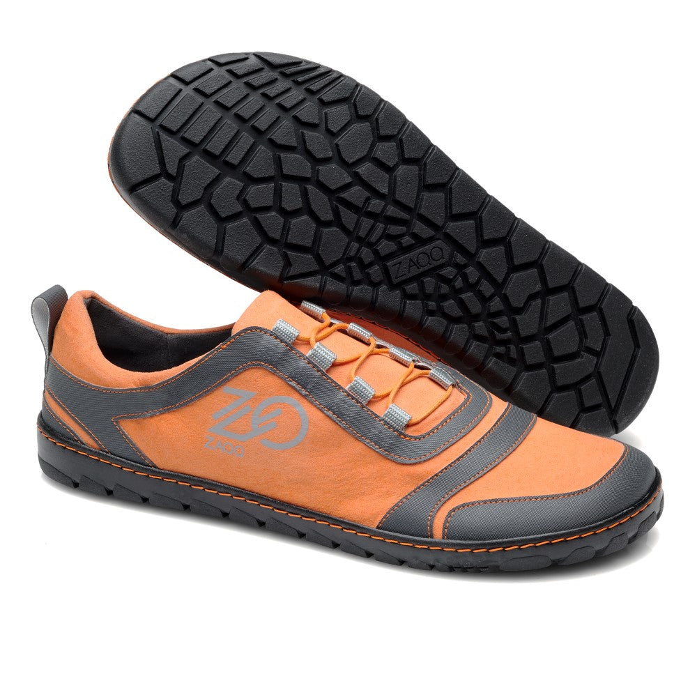 Der SQILL Orange von ZAQQ ist ein Sportschuh in orange-grauem Design mit strukturierter schwarzer Sohle. Er verfügt über ein einzigartiges Schnürmuster und zeigt das Logo der Marke an der Seite. Ein Schuh steht aufrecht, während der andere sein aufwendiges Sohlendesign hervorhebt.