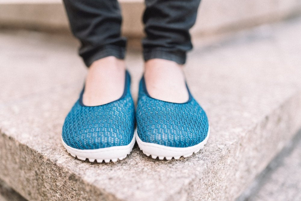 Eine Person in den Slippern SUQAR Mare von ZAQQ, mit blauem Strukturdesign aus Baumwoll-Strickgewebe und weißer Sohle, steht auf Steinstufen. Sie trägt eine schwarze Hose.