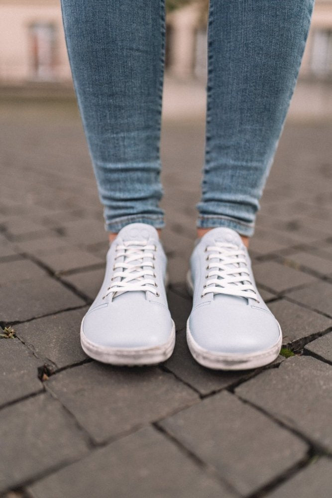 Eine Person in den TAQQ Nappa Lightblue Sneakers mit flexibler Sohle von ZAQQ und blauen Skinny Jeans steht auf einem Kopfsteinpflaster. Die sauber glänzenden Schnürschuhe harmonieren perfekt mit der eng sitzenden Jeans vor dem leicht unscharfen Hintergrund.