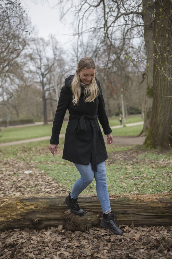 Eine Frau in schwarzem Mantel und Jeans balanciert auf einem umgestürzten Baumstamm im Park. Ihre TREQ Black Winter Waterproof-Stiefel von ZAQQ sorgen für einen sicheren Stand. Die Bäume mit spärlichem Laub und Gras im Hintergrund lassen auf einen kühlen, bewölkten Tag schließen.