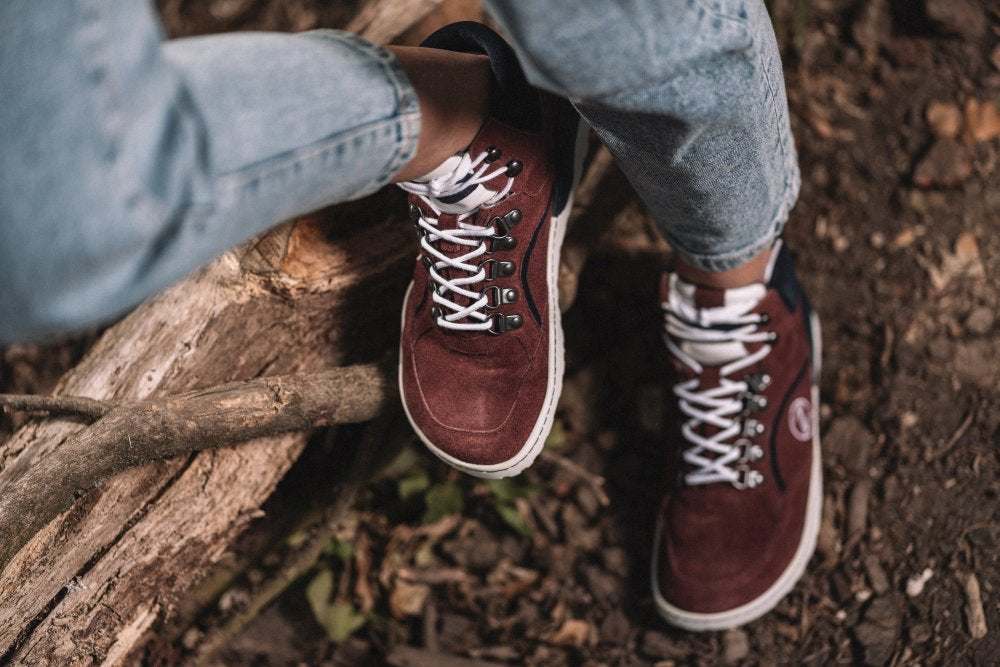 In den TREQ Red Waterproof Sneakers von ZAQQ sitzt eine Person auf einem umgestürzten Baumstamm und zeigt damit die ideale Mischung aus Stil und Funktionalität, die für Outdoor-Ausrüstung typisch ist. In hellblauen Jeans gekleidet, entspannt sie sich zwischen Dreck und verstreuten Blättern und schätzt den wasserdichten Komfort, den diese Schuhe bieten.