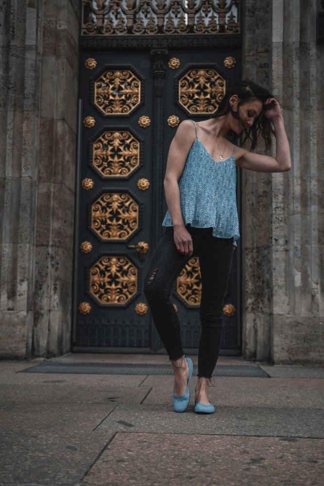 Eine Frau mit langen Haaren steht vor einer verzierten Tür mit goldenen Details. Sie trägt das ärmellose TWIST Light Blue Top von ZAQQ, kombiniert mit schwarzen Jeans und flexiblen Schuhen. Sie blickt nach unten und berührt sanft ihr Haar in einer Umgebung, die eine elegante, stimmungsvolle Atmosphäre ausstrahlt.