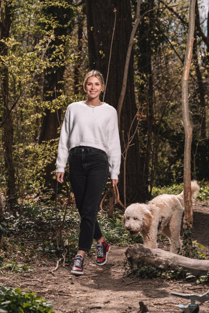 Eine Frau in weißem Pullover und schwarzer Hose führt einen flauschigen Hund an der Leine durch einen sonnenbeschienenen Wald. Mit farbenfrohen VAQATION Red Waterproof Sneakers von ZAQQ lächelt sie, während sie den von Bäumen und Grün umgebenen Waldweg entlangschlendert.