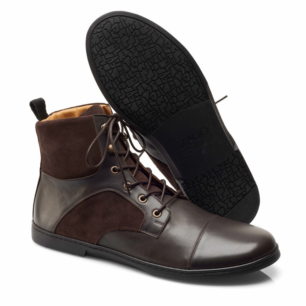 Ein Paar stylische, dunkelbraune Stiefeletten aus Leder und Wildleder aus der YORQ Brown-Kollektion von ZAQQ mit schwarzen Gummisohlen. Ein Stiefel steht aufrecht, während der andere geneigt ist und die strukturierte Sohle mit einem geometrischen Muster zur Schau stellt.